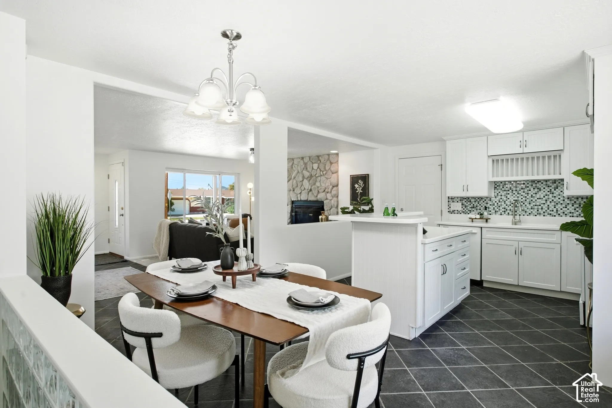 Open-concept living area featuring a dining space with a chandelier, a kitchen with white cabinetry and mosaic tile backsplash, and dark tile flooring