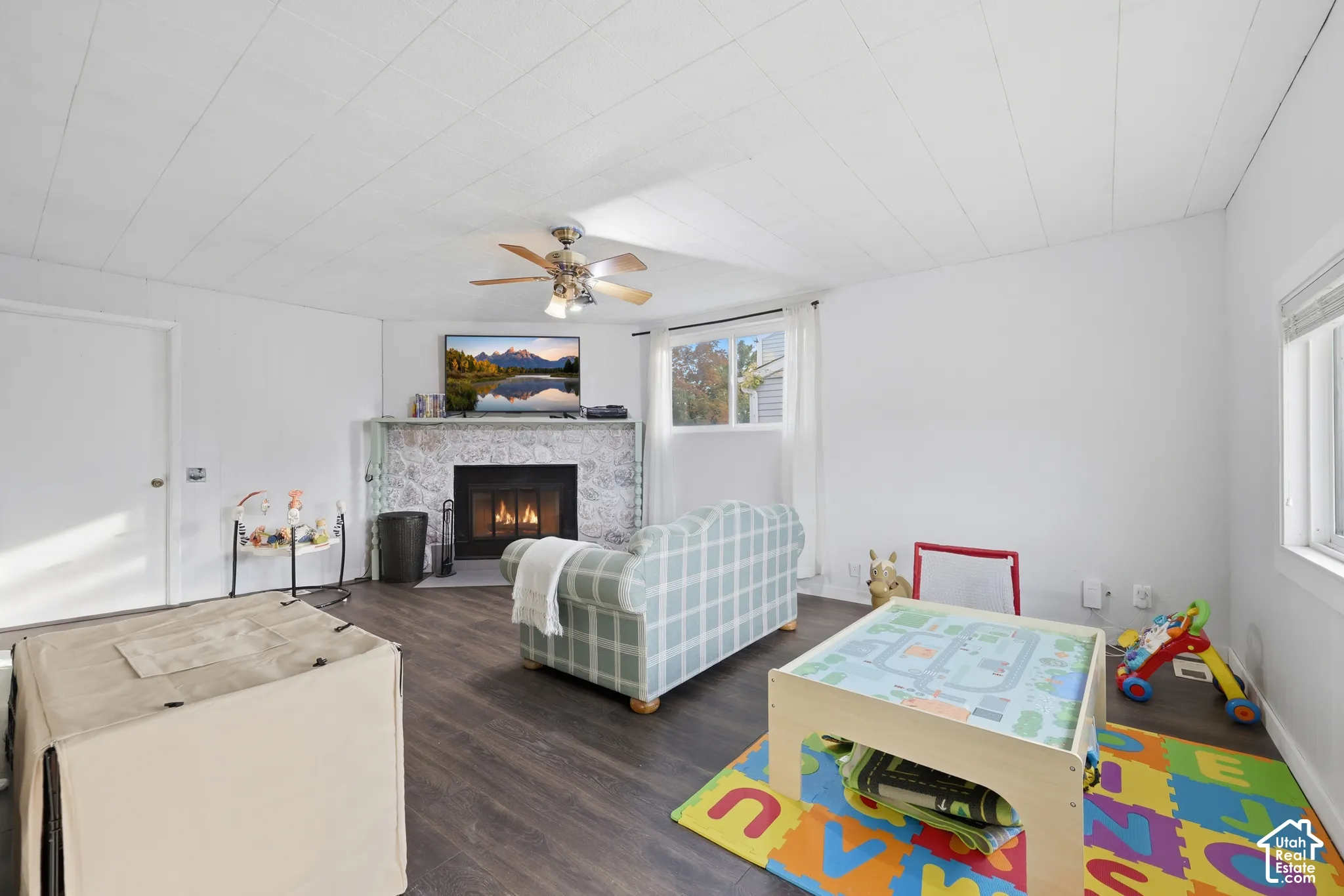 White paneled ceiling, ceiling fan with light fixture, stone-clad fireplace, dark wood-finish flooring, and white window blinds