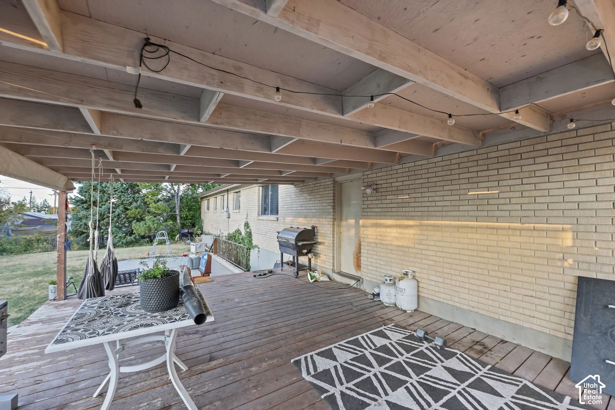 Covered wooden deck with exposed beam ceiling, string lighting, and light-colored brick wall