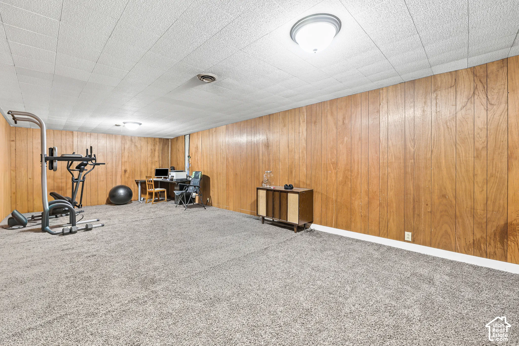 Expansive room featuring wood-panel walls, neutral carpet flooring, and a drop ceiling with recessed lighting