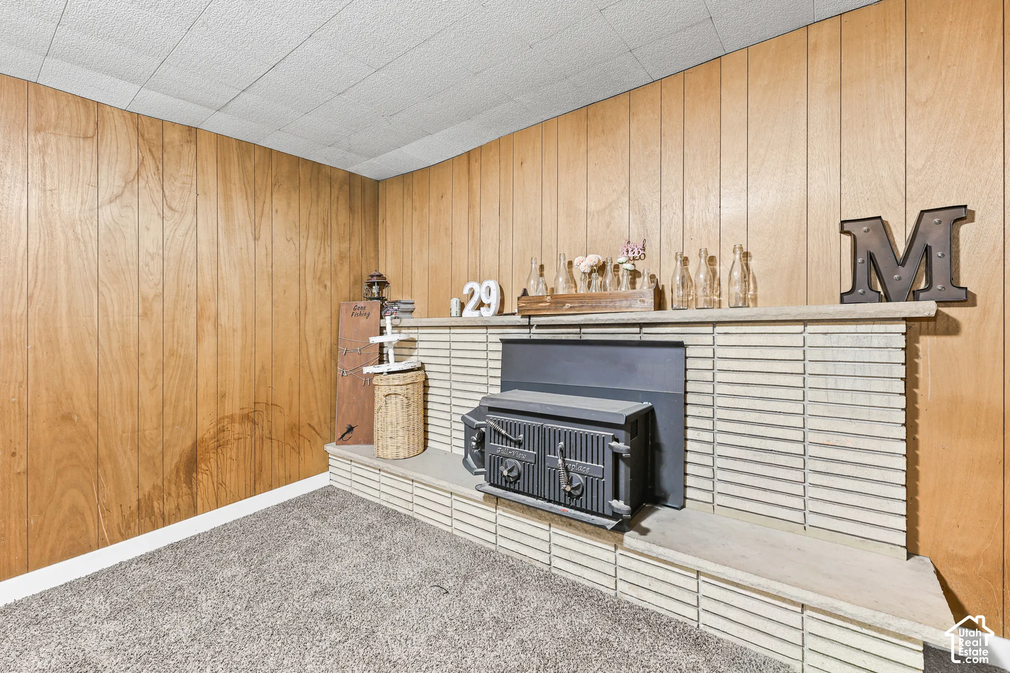 Wood paneling walls, textured ceiling, stone fireplace surround, dark metal firebox insert, and gray carpet