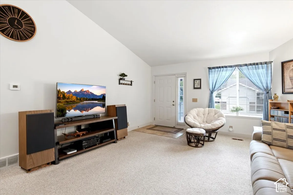 Vaulted ceiling, white walls, carpeted flooring, paneled entry door, and a large window with blinds