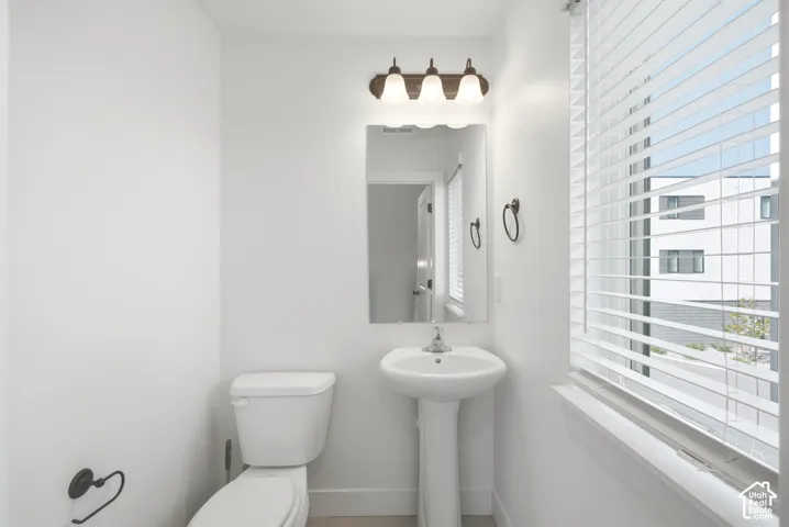 Pedestal sink with chrome faucet, wall-mounted mirror, three-light vanity fixture, white toilet, and window with horizontal blinds