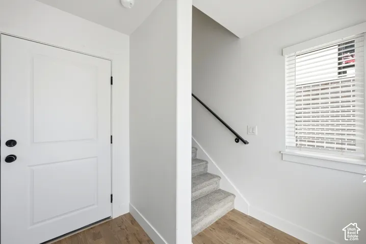 Entryway featuring a white paneled door with matte black hardware, wood-finish flooring, a carpeted staircase with a black handrail, and a window with white blinds