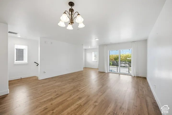 Spacious room with wood-finish flooring, white walls, a multi-light chandelier, and sliding glass doors opening to a balcony with a black metal railing