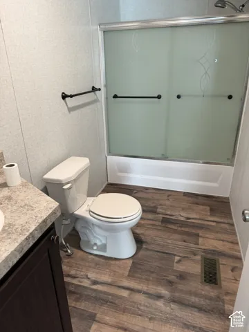 Wood-finish flooring with a white toilet, brown vanity, and a shower-over-tub with frosted glass sliding doors