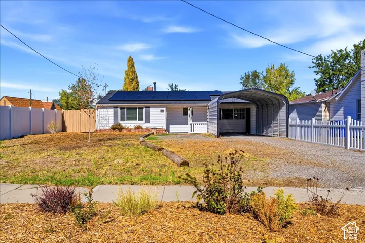 White exterior with dark-paneled solar array, side carport, gravel driveway, and a partially fenced yard