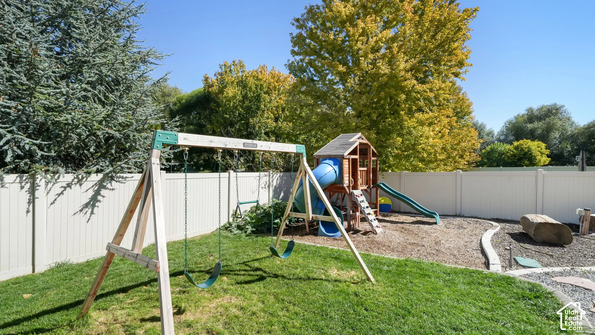 Fenced backyard featuring a manicured lawn, dedicated play area with wood chips, and mature trees providing shade