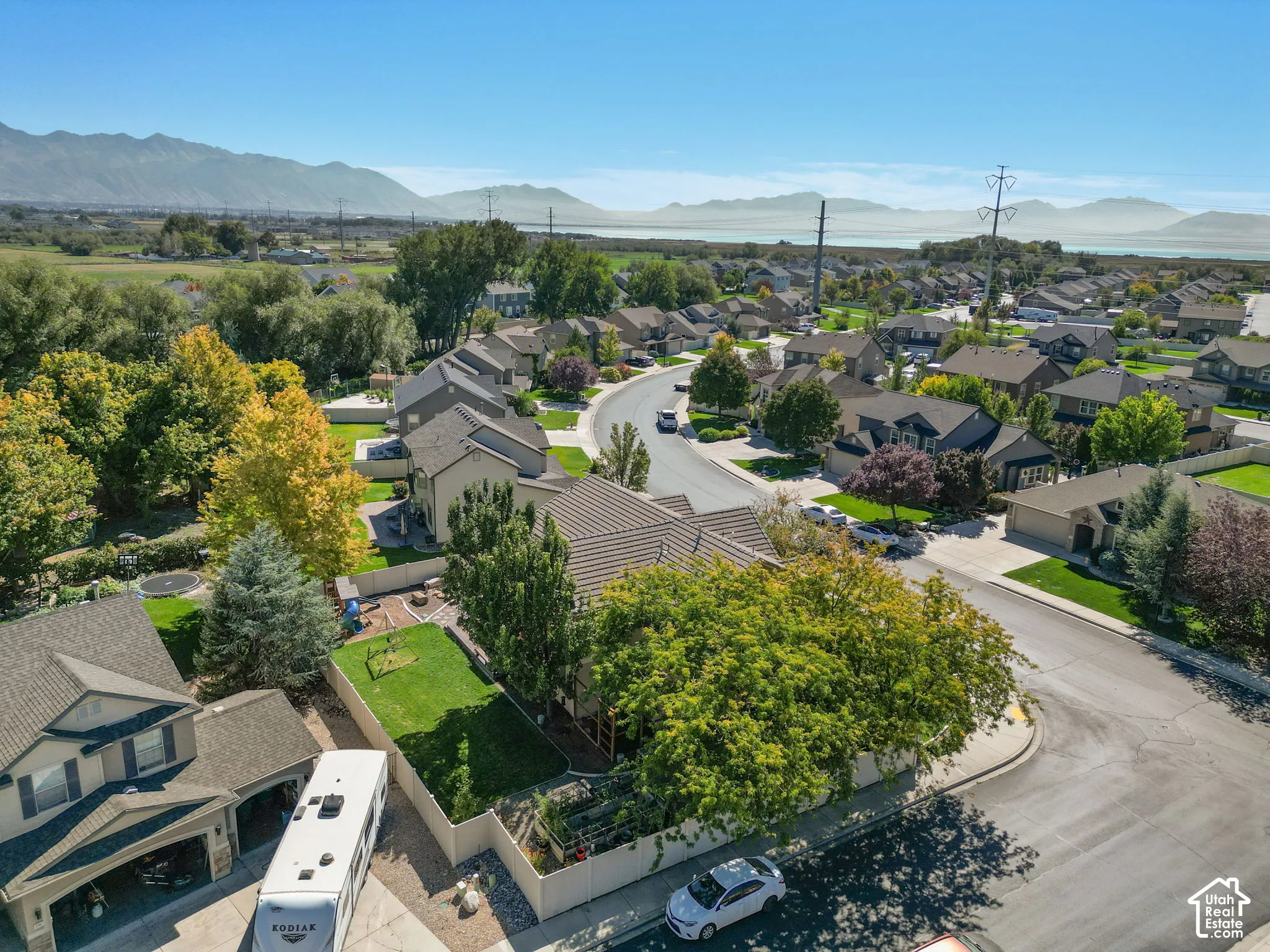 Residential neighborhood with mature trees and paved streets, bordered by expansive green fields and distant mountains