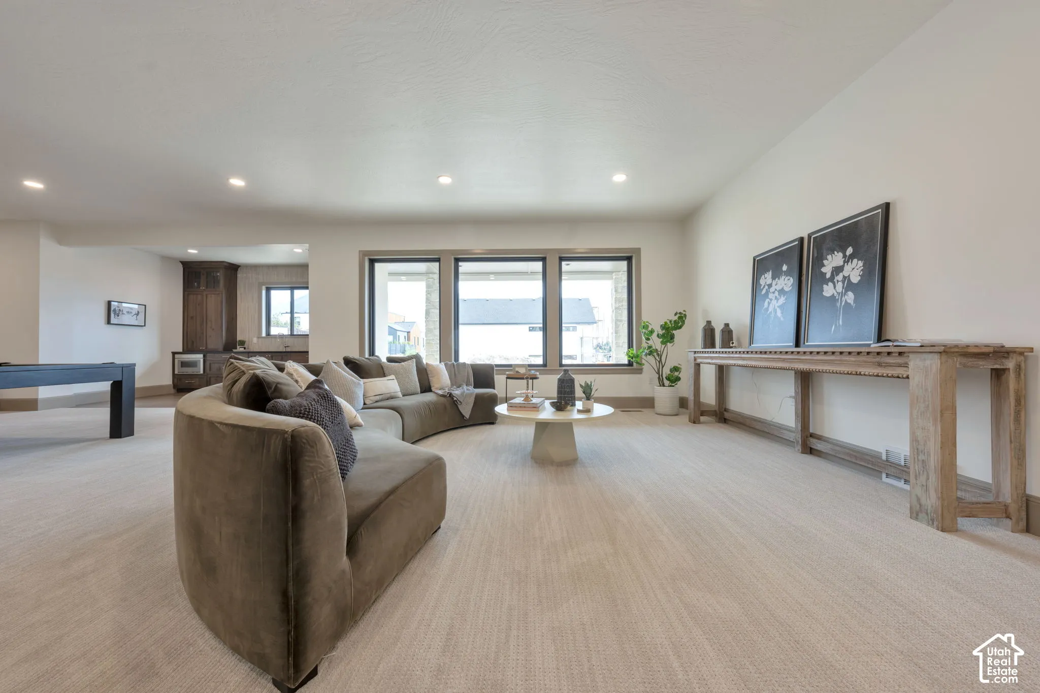 Recessed lighting illuminates a spacious room featuring large windows with dark trim, light-toned carpet, and a built-in wet bar with dark wood cabinetry