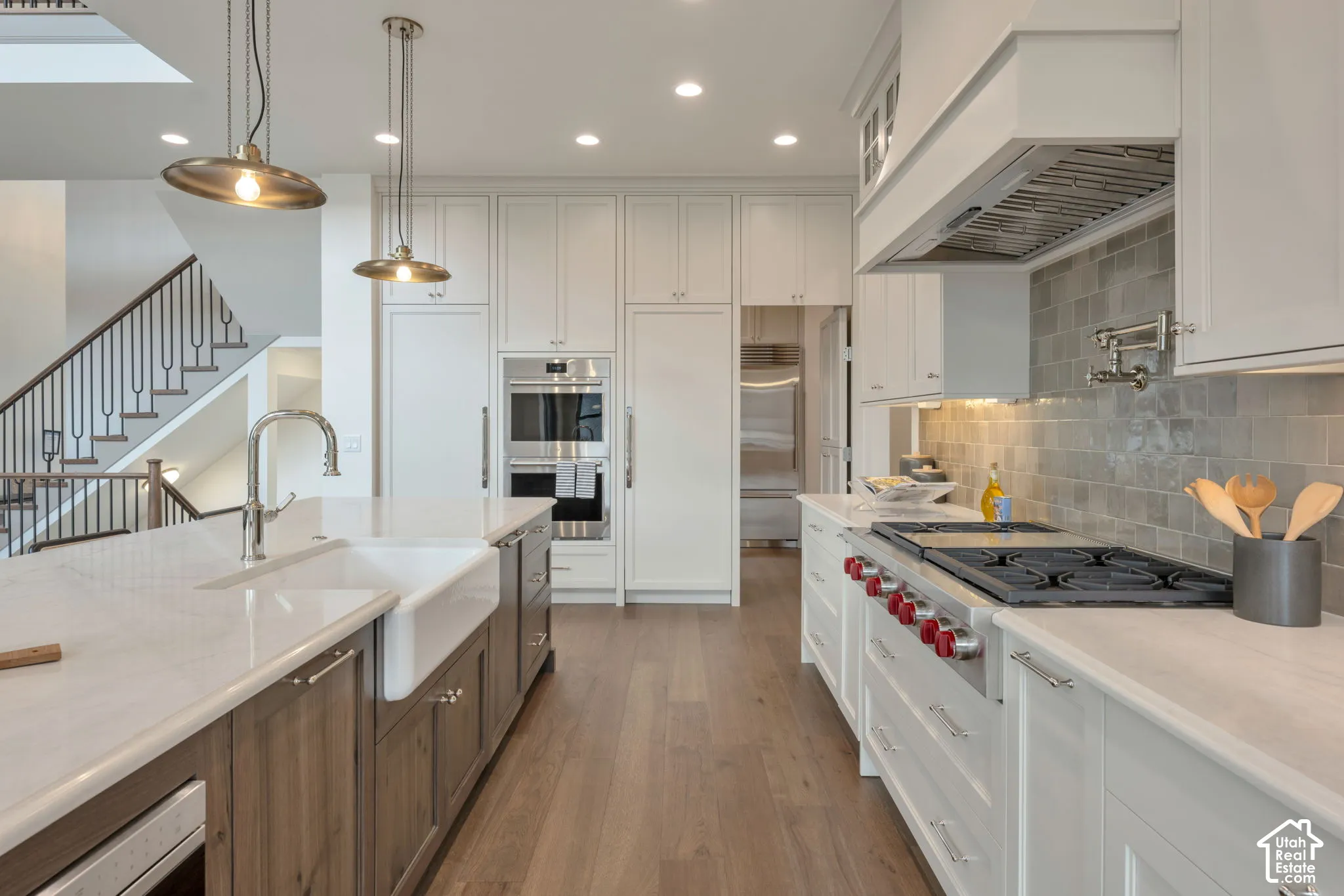 Gourmet kitchen featuring a large island with a farmhouse sink, integrated stainless steel appliances, custom cabinetry, a tile backsplash, and wood-finish flooring