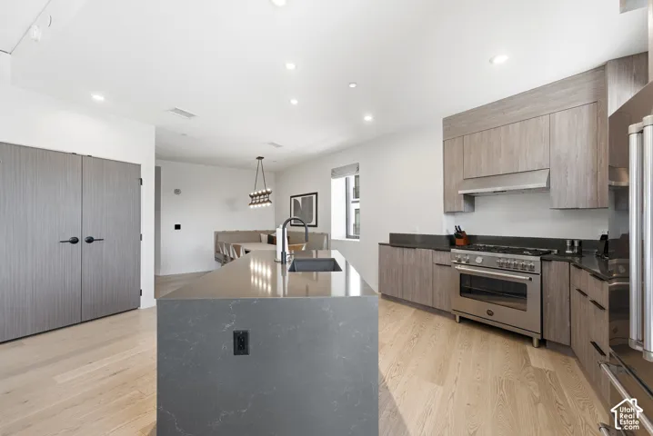 Modern kitchen featuring light wood-finish flooring, a large island with a built-in sink, stainless steel range with overhead hood, and light wood cabinetry with dark countertops