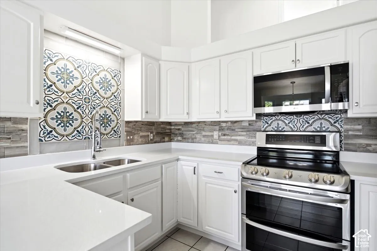 White cabinetry with silver hardware, dual basin stainless steel sink, patterned tile backsplash, stainless steel appliances, and light-toned countertops