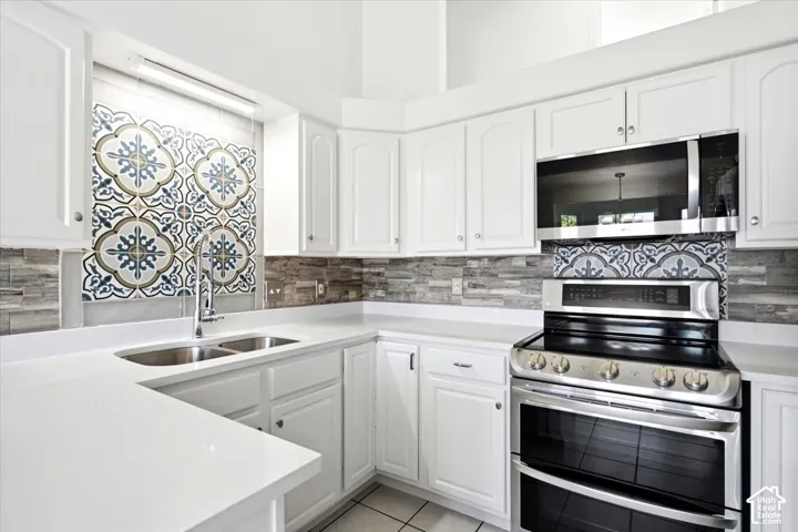 White cabinetry with silver hardware, dual basin stainless steel sink, patterned tile backsplash, stainless steel appliances, and light-toned countertops