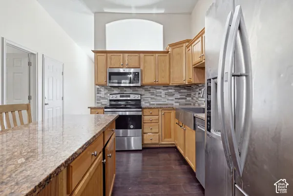 Kitchen featuring wood cabinetry, stone-finish countertops, stainless steel appliances including a farmhouse-style sink, and a multi-tone tile backsplash with wood-finish flooring