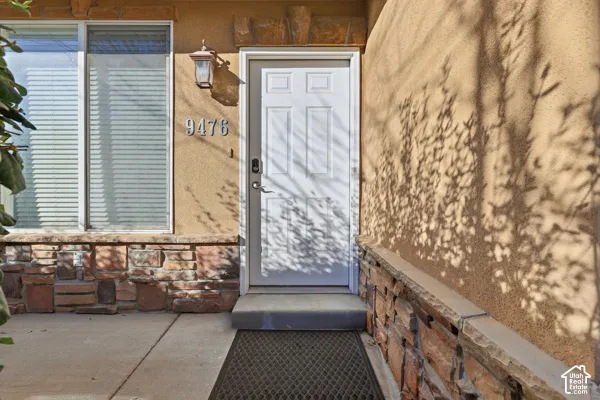 Stucco exterior featuring stacked stone veneer, a six-panel entry door, concrete threshold, and mounted wall lighting