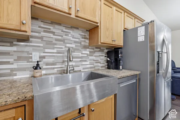 Stainless steel farmhouse sink featuring a high-arc faucet, complemented by light wood cabinetry, stone-finish countertops, and a multi-tonal linear tile backsplash