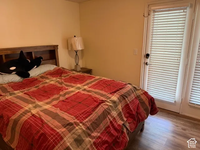 Comfortable bedroom featuring warm wood-finish flooring, a wood headboard bed frame, bedside table with attached lamp, and glass door with louvered shutters providing natural light and access to outdoor space