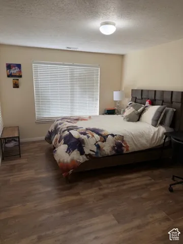 Spacious bedroom featuring a wood-finish plank floor, large picture window with horizontal blinds providing natural light, recessed ceiling lighting, and a padded upholstered bed frame