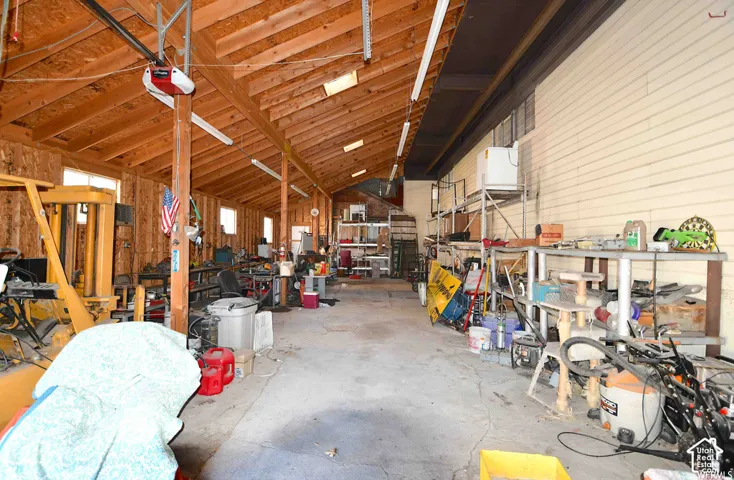 Spacious garage featuring exposed wood beams, concrete flooring, ample natural light from multiple windows, and extensive shelving for storage