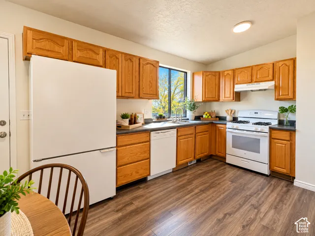 Kitchen featuring wood-finish flooring, wood cabinetry, dark countertops, a window over the sink, and white appliances including a refrigerator, dishwasher, and range