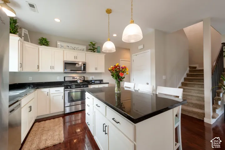 Kitchen featuring white cabinetry, dark countertops, stainless steel appliances, a central island with pendant lighting, and wood-finish flooring