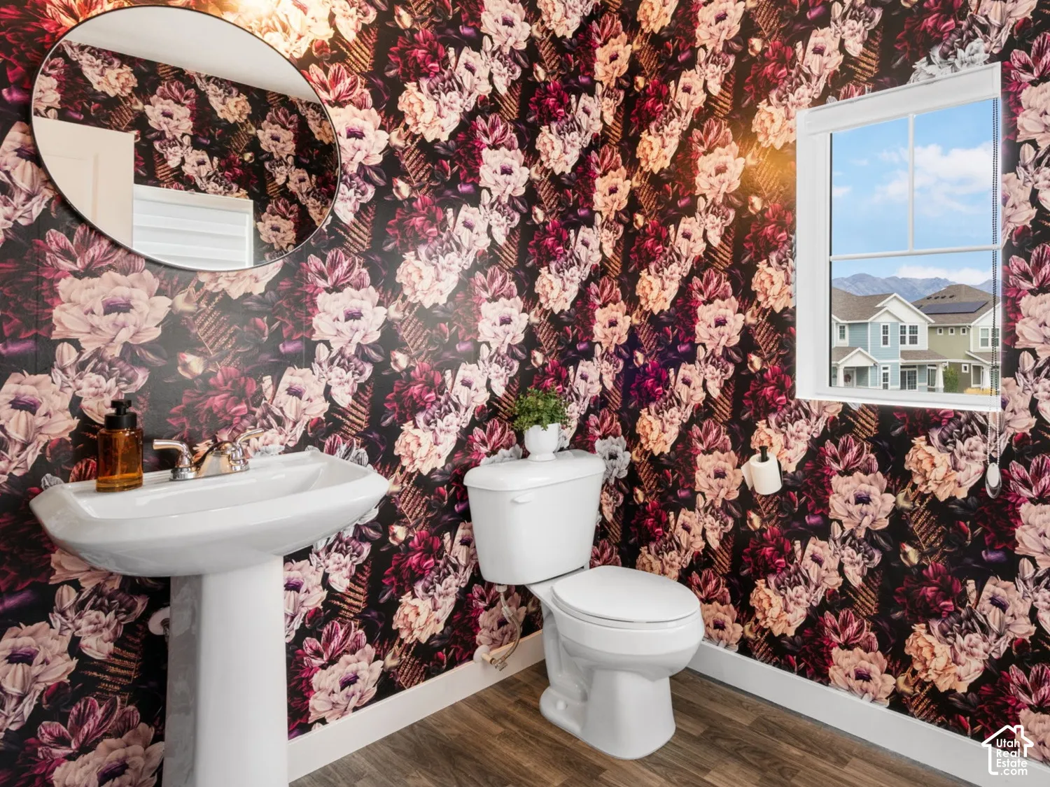Pedestal sink, round wall mirror, decorative floral wallpaper, wood-finish flooring, and a single window