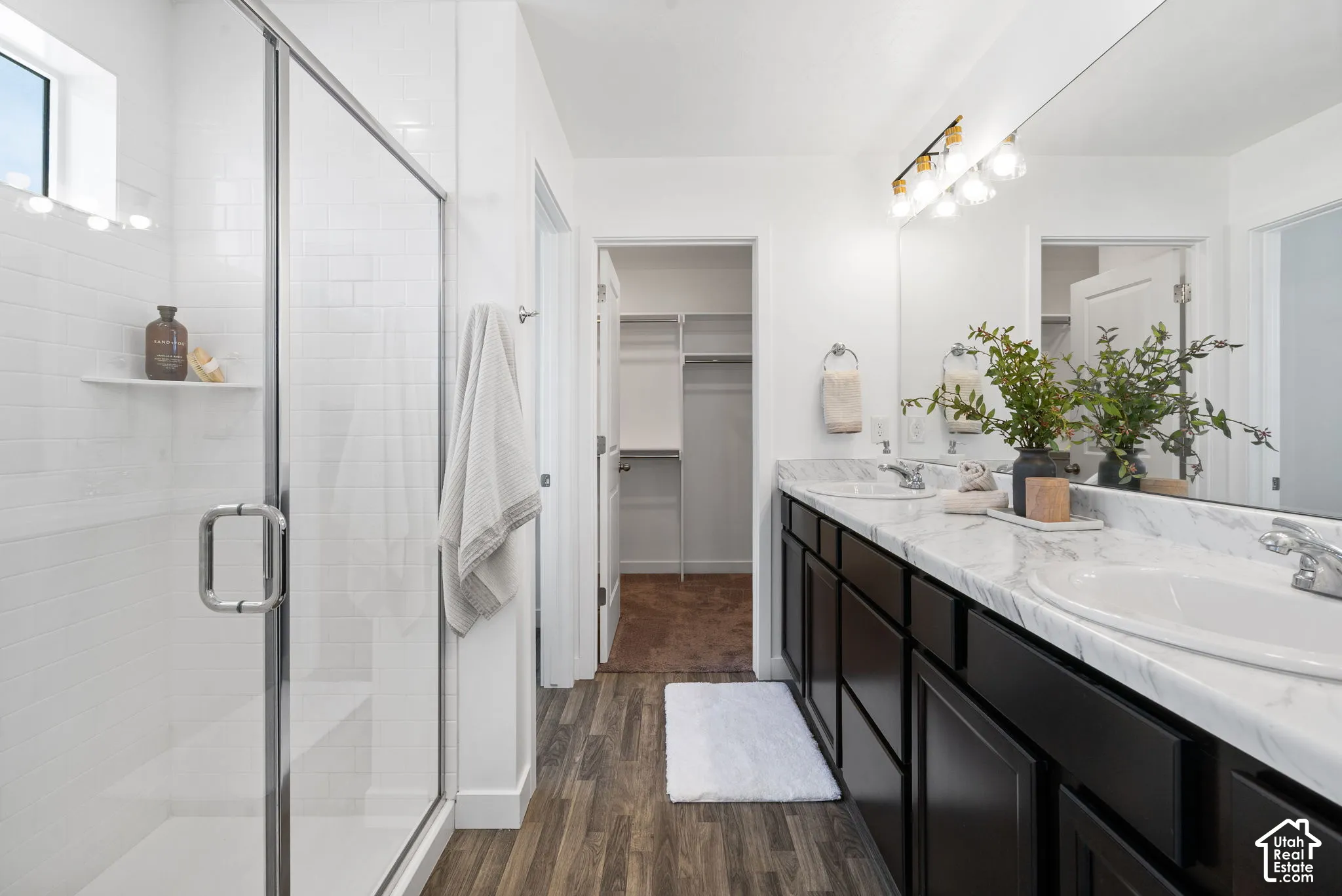 Enclosed glass shower with white subway tile surround, dual vanity with undermount sinks, expansive mirror, dark wood-finish cabinetry, and wood-finish flooring