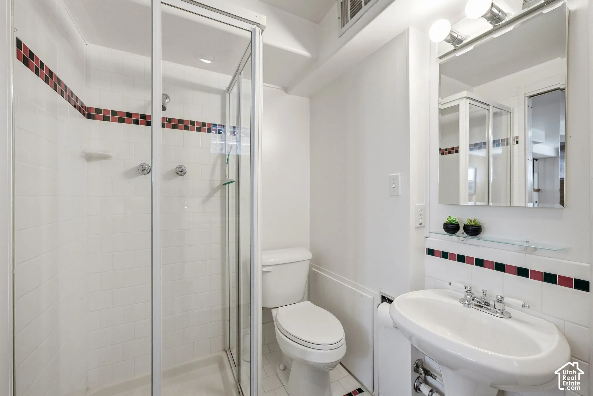 Pedestal sink with chrome fixtures, mirrored medicine cabinet, and a glass-enclosed shower with subway tile surround and decorative tile accent
