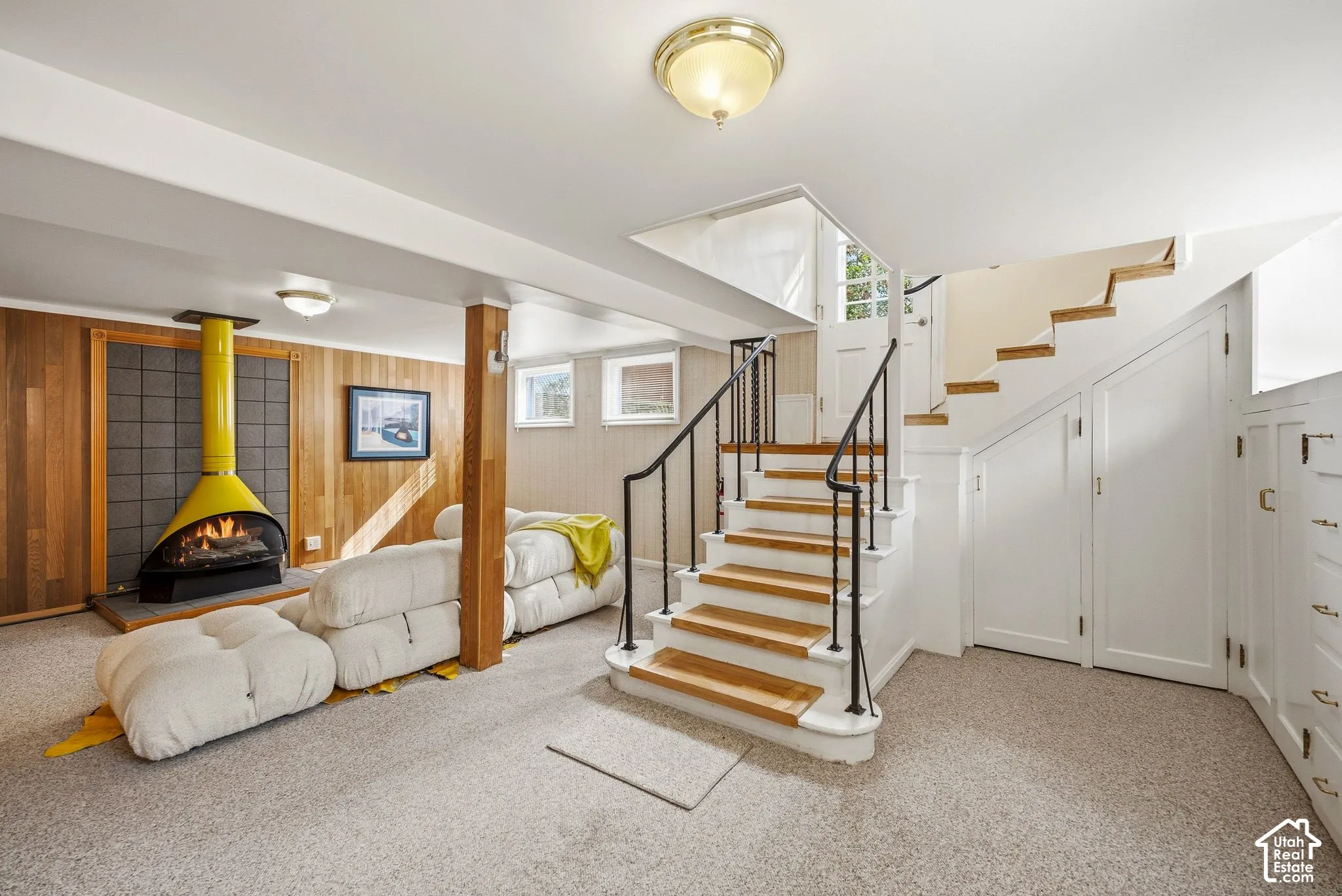 Wood-paneled wall with a modern freestanding fireplace, carpeted flooring, and a staircase with wood treads and a black metal railing. Extensive storage