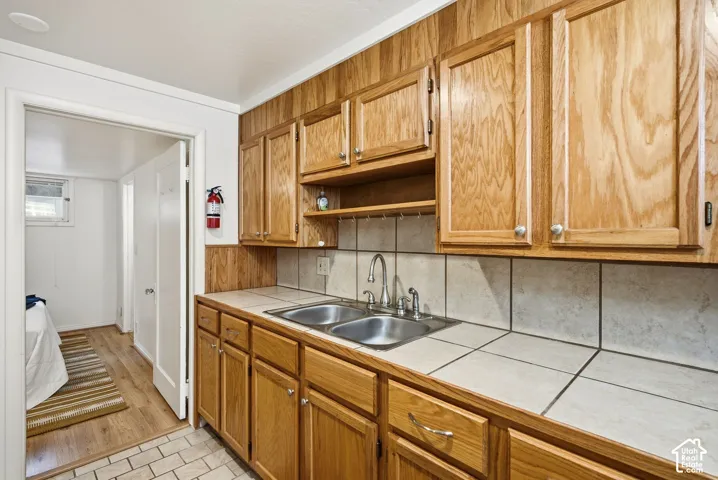 Mother in Law appt kitchen featuring warm wood cabinetry with ample storage, stainless steel sink with chrome faucet, ceramic tile backsplash, and tile flooring throughout. Door to bedroom