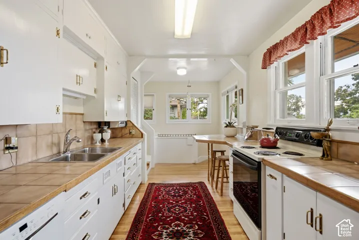 White cabinetry with brass-finish hardware, tile countertops, a stainless steel double basin sink, and hardwood flooring