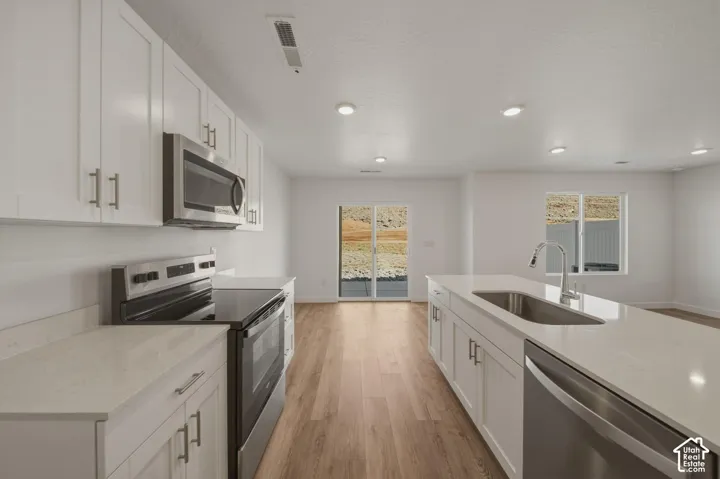 Kitchen featuring white cabinetry, stainless steel appliances, light-toned countertops, a kitchen island with an undermount sink, and wood-finish flooring