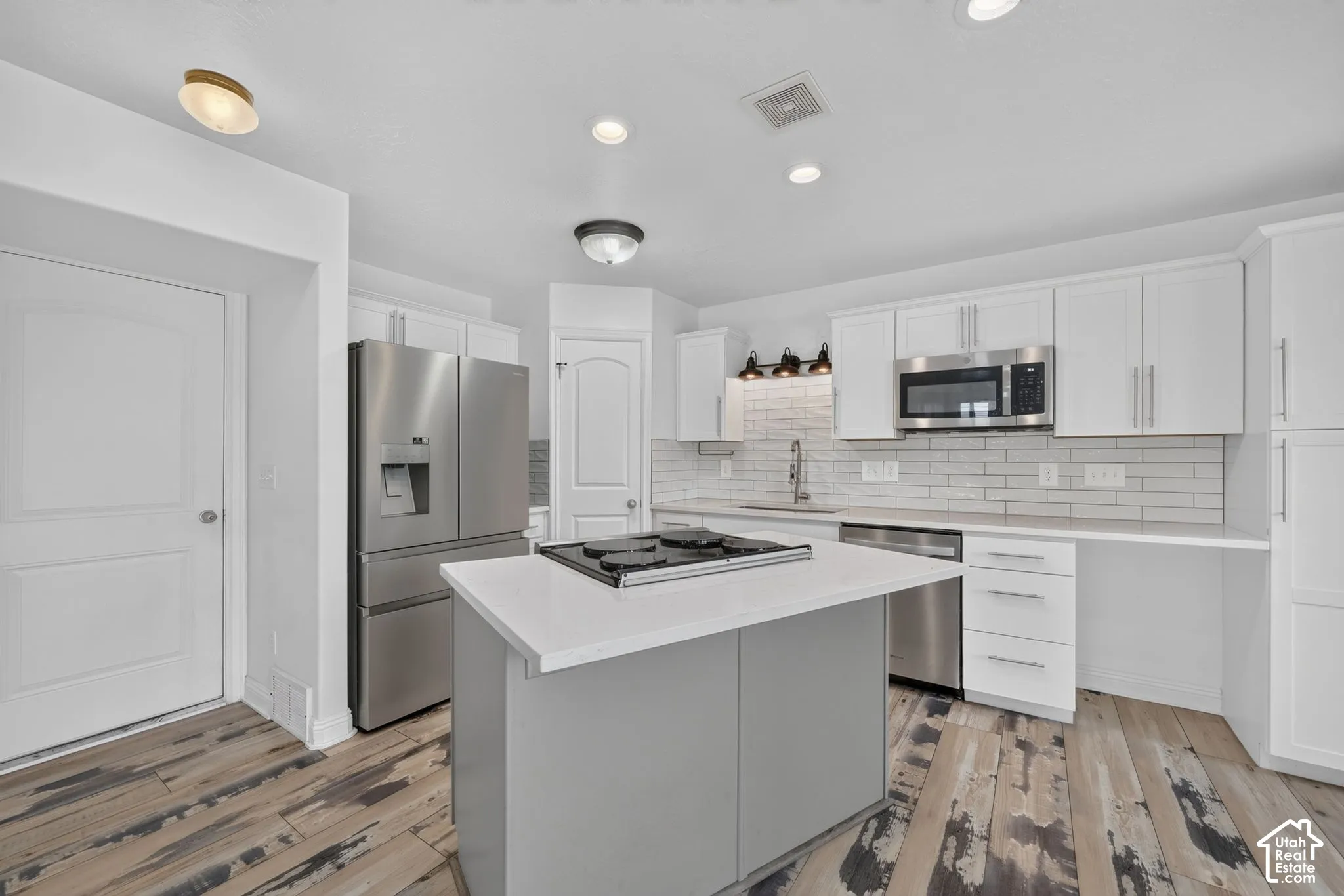 Kitchen featuring an island with a built-in cooktop, white cabinetry, stainless steel appliances, white subway tile backsplash, and wood-finish flooring