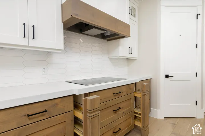 White flat-panel cabinetry with black hardware, wood-finish base cabinetry, light-toned countertops, white picket-patterned backsplash, and integrated cooktop