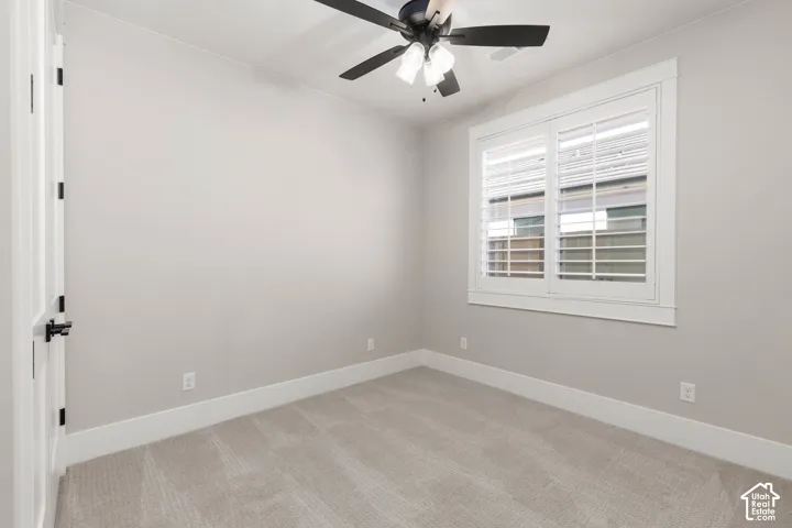 Bright, light-filled bedroom showcasing neutral wall finishes, plush carpet flooring, a ceiling fan with integrated lighting, plantation shutters framing the window, and white trim detailing throughout