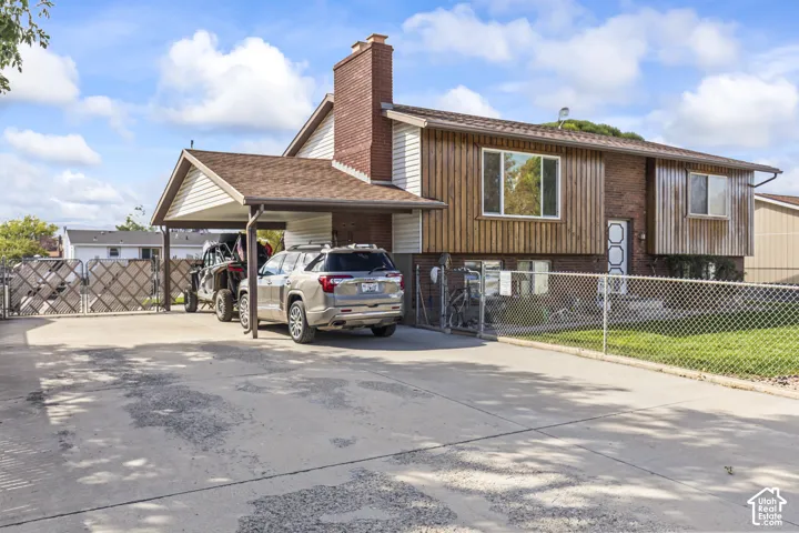 Brick and wood siding exterior with a covered carport, paved driveway, chain-link fencing, and a red brick chimney feature