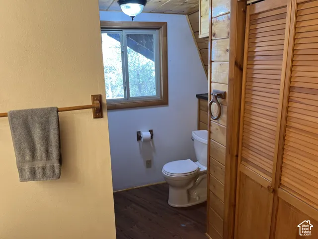 Wood-finish flooring, white toilet, wood-paneled wall, wood-framed window, and wood louvered doors