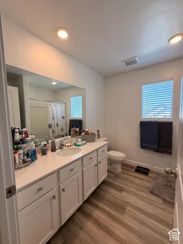 Dual vanity with integrated sinks, white cabinetry, a large wall-mounted mirror, wood-finish flooring, and a window with blinds