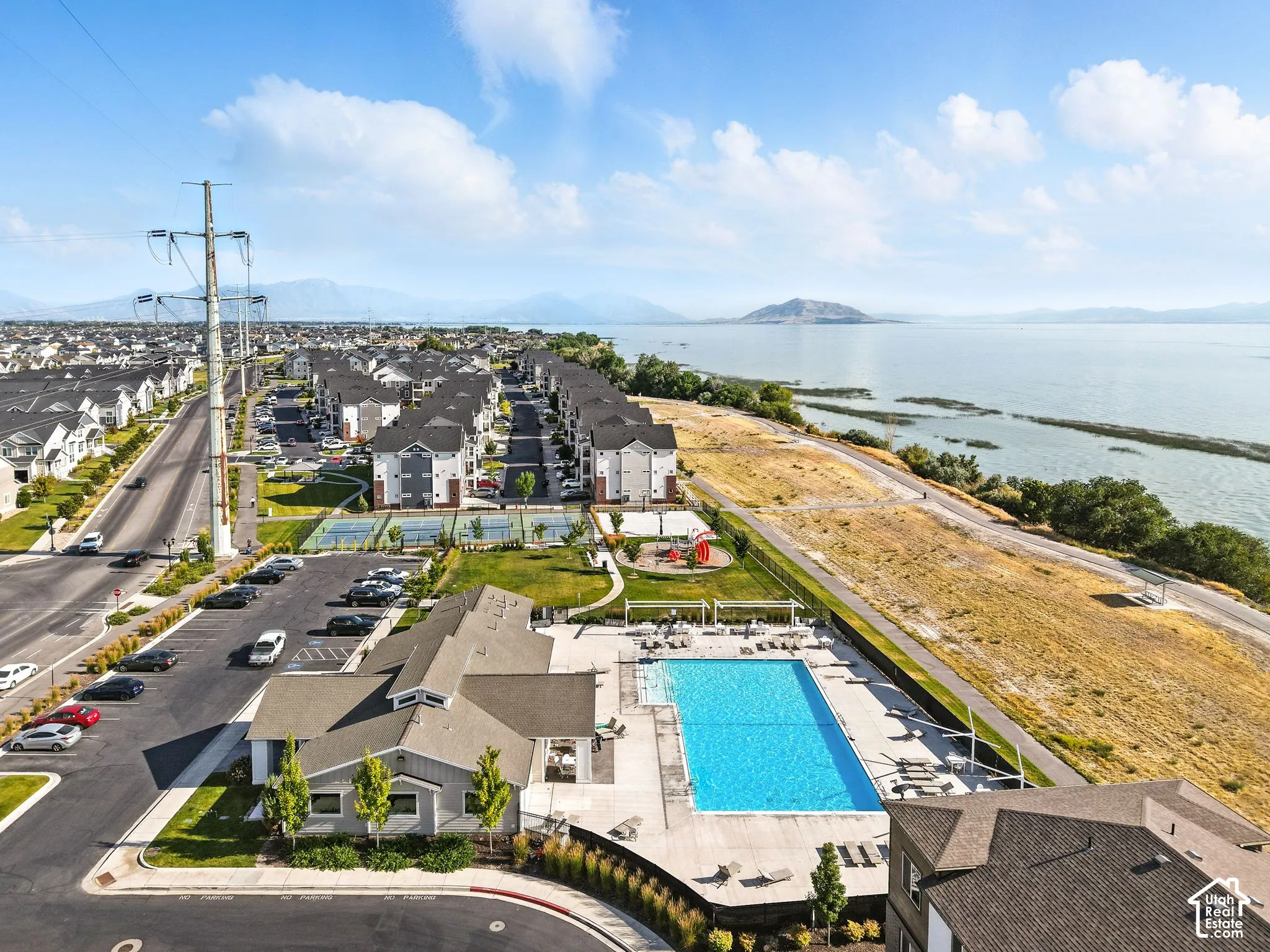 Expansive outdoor swimming pool with surrounding lounge areas, integrated tennis courts, playground area, and a large body of water in the background