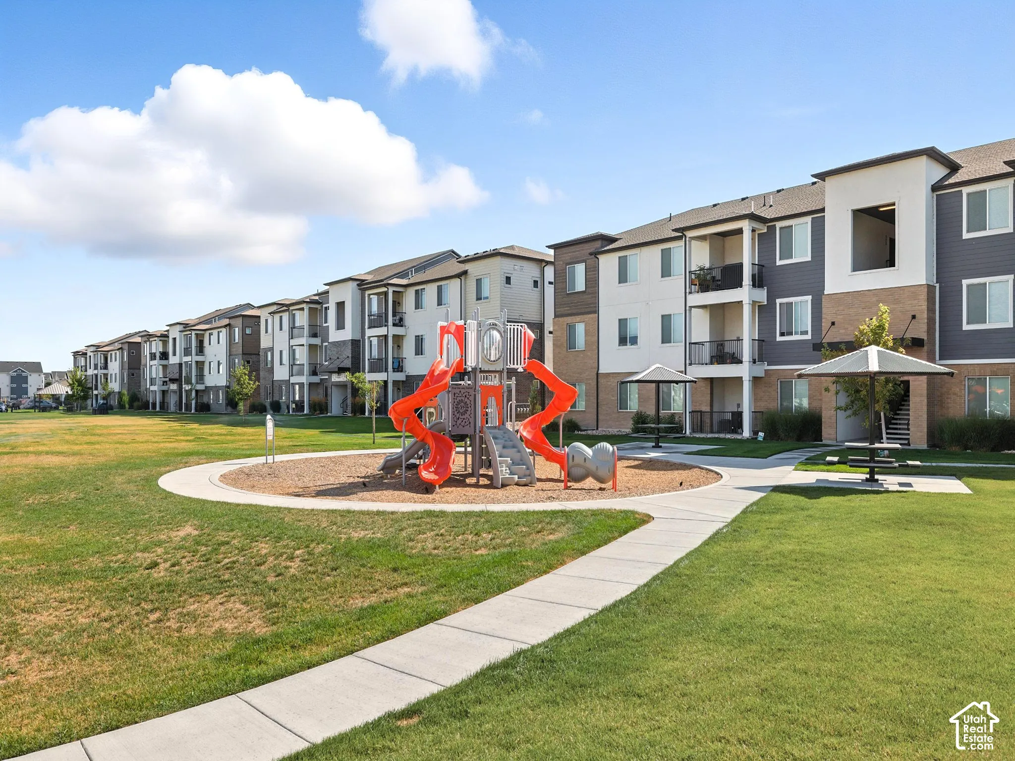 Expansive grassy area featuring a playground, curved concrete pathway, and covered picnic area with seating