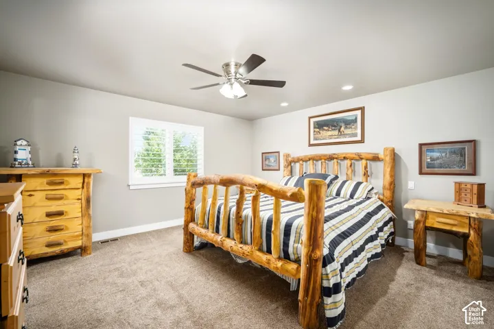 Spacious bedroom featuring warm wood-finish bed frame, neutral carpeting throughout, ceiling fan with integrated lighting, and large window providing natural light
