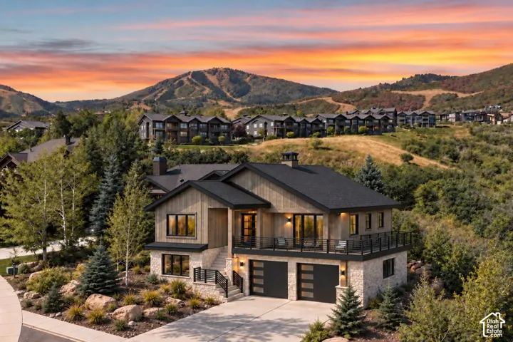 Two-story mountain residence featuring vertical wood siding, stone masonry base, a wraparound metal-railing deck, and a two-car garage with horizontal window panes, set against a backdrop of mountainous terrain