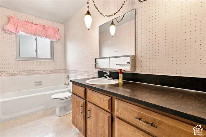Wood-finish vanity with a single basin sink and dark countertop, framed mirror with integrated lighting, and a white bathtub with tile surround