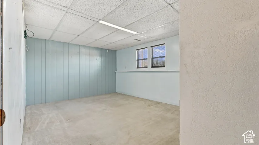 Room featuring a drop ceiling with integrated fluorescent lighting, wood-paneled walls, carpeted flooring, and dual-pane windows