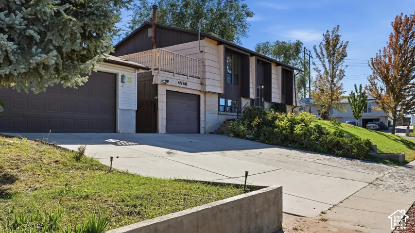 Split-level exterior featuring a one-car and two-car garage, upper-level wood deck, concrete driveway, and concrete retaining wall