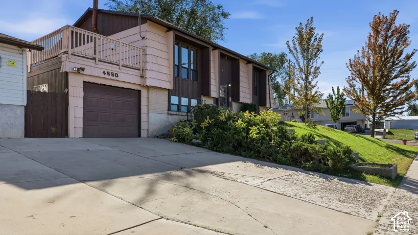 Split-level exterior featuring a concrete driveway, attached garage, wood-trimmed upper deck, and landscaped front yard