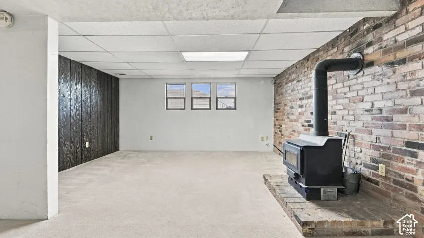 Finished space featuring a brick accent wall, wood-burning stove on a raised hearth, wood paneling, and drop ceiling