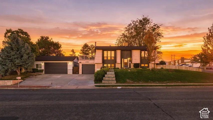 Split-level exterior featuring concrete driveway, two-car garage, upper-level balcony, and landscaped lawn with stone stairs