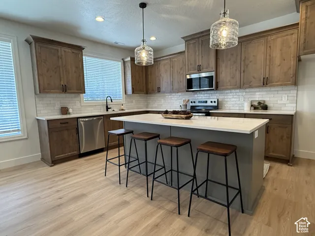 Kitchen featuring wood-finish cabinetry, stainless steel appliances, a white subway tile backsplash, light-colored countertops, and pendant lighting